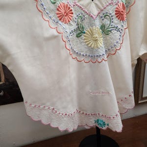 May include: Cream-colored embroidered blouse with a V-neckline. The blouse features colorful floral embroidery in shades of pink, yellow, and green. The text "Costa Rica" is embroidered on the lower part of the blouse.