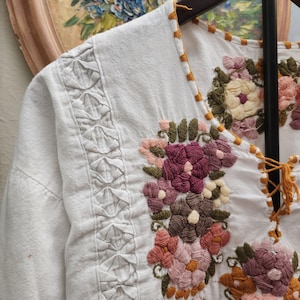 Puede incluir: Blusa blanca bordada con dise&ntilde;os florales en tonos rosa, morado y marr&oacute;n. La blusa presenta costuras detalladas a lo largo del escote y las mangas, y un frente con cordones. El bordado a&ntilde;ade un toque vintage a la prenda.