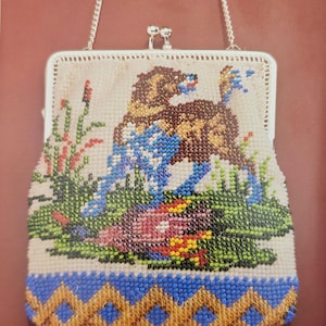 May include: A rectangular beaded handbag featuring a multi-colored needlepoint design of a brown dog in a grassy field. The bag has a silver-toned metal frame with a kiss-lock closure and a thin silver chain strap. The base has a blue and gold pattern.