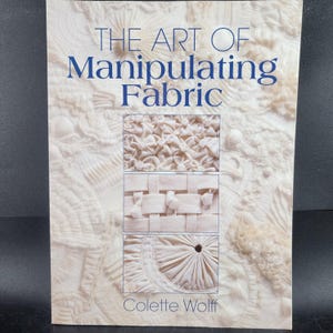 May include: A book titled "THE ART OF Manipulating Fabric" by Colette Wolff. The cover features a cream-colored background with three fabric manipulation examples: gathered, woven, and pleated. The title is in blue.