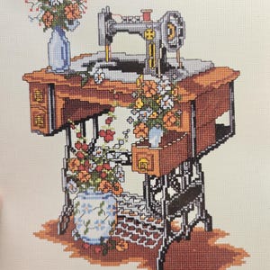 May include: A cross-stitch pattern featuring a vintage sewing machine on a wooden table with drawers. The design includes blue vases filled with orange and yellow flowers, set against a light fabric background with a brown floor shadow.