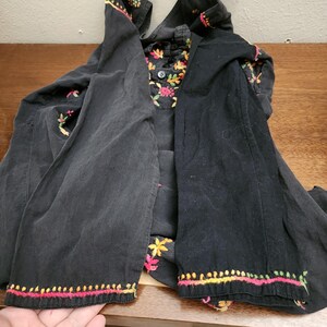 Peut inclure: Veste noire brod&eacute;e avec des accents floraux color&eacute;s. La veste pr&eacute;sente un col et des poignets avec des broderies complexes dans les tons de rose, de jaune et de vert. La doublure int&eacute;rieure de la veste a un motif floral.