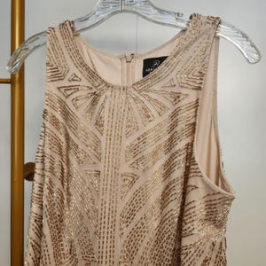 May include: A sleeveless beige dress featuring an intricate geometric pattern of metallic gold beads across the front. The garment has a round neckline, a central back zipper, and a label inside the collar that reads Adrianna Papell.