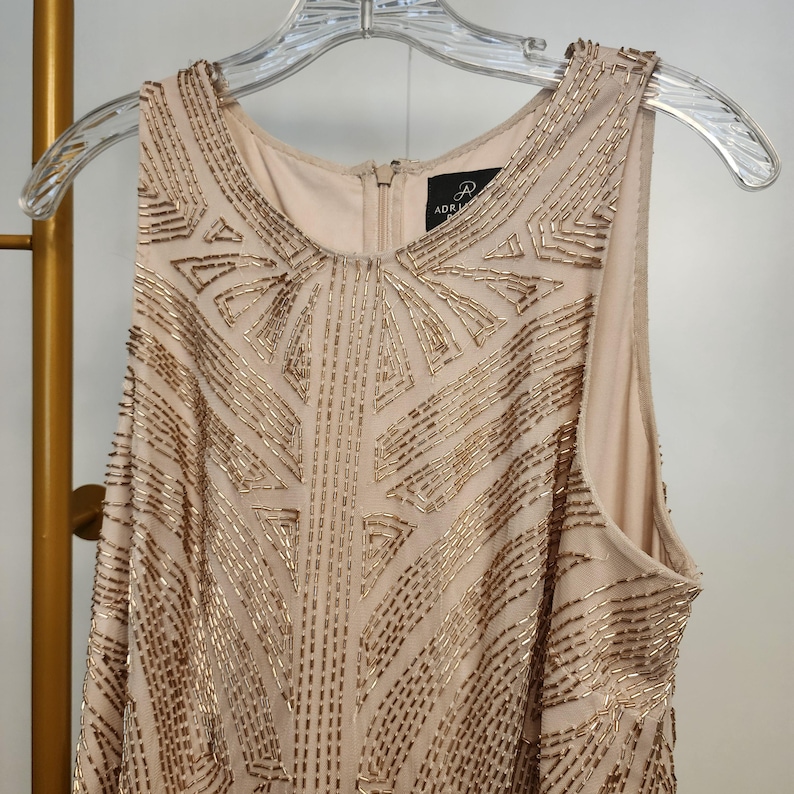 May include: A sleeveless beige dress featuring an intricate geometric pattern of metallic gold beads across the front. The garment has a round neckline, a central back zipper, and a label inside the collar that reads Adrianna Papell.