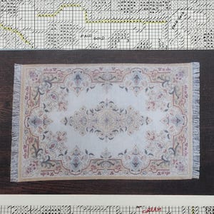 May include: A miniature rectangular rug with a cream-colored central field and an ornate floral pattern in shades of beige, pink, and blue. The rug has a fringed border and is likely for a dollhouse or miniature setting.