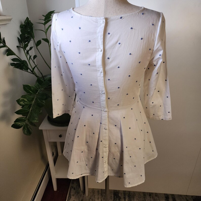 May include: A white dress with a blue polka dot pattern and a button-up back. The dress has a flared skirt and three-quarter sleeves.