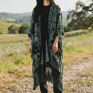 May include: A sage green open-front kimono featuring a black velvet floral pattern throughout the fabric. The garment has long, loose sleeves, a straight hemline, and is finished with long, matching sage green fringe along the bottom edge.