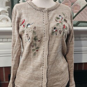 May include: Tan cardigan sweater with embroidered birds and pine branches. The long-sleeved sweater has a button-up front and a round neckline. The embroidery features red, black, white, and green accents. The sweater is displayed on a mannequin.