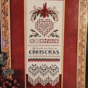 May include: A framed Christmas cross-stitch design with a red border. The design features a heart, holly, and the words "All hearts are home on CHRISTMAS". The frame is gold.
