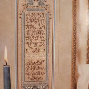 May include: A framed cross-stitch sampler featuring the alphabet in uppercase and lowercase letters. The sampler is stitched in brown thread on a beige fabric, with decorative borders and floral accents. The frame is a light beige color.