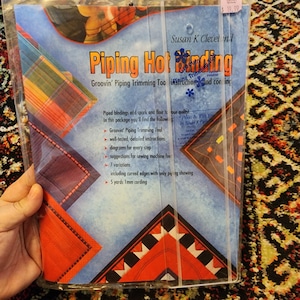 May include: A package for "Piping Hot Binding" by Susan K Cleveland. The package includes a "Groovin' Piping Trimming Tool", detailed instructions, diagrams, and 5 yards of 1mm cording. The package has a blue background with colorful geometric designs.