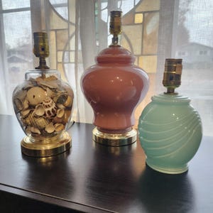 May include: Three decorative table lamps. One is a clear glass jar filled with seashells, another is a peach-colored ceramic lamp, and the third is a light teal lamp with a swirled design. All have brass bases and lamp holders.