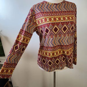 May include: Long-sleeved shirt with a geometric pattern in shades of red, yellow, and blue. The pattern is reminiscent of Native American designs.
