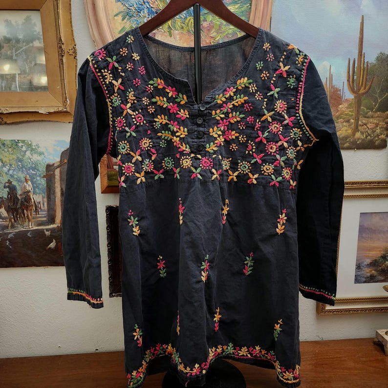 Peut inclure: Une tunique noire &agrave; manches longues avec des broderies florales color&eacute;es. La broderie pr&eacute;sente des fleurs et des feuilles roses, jaunes, vertes et oranges. La tunique a un col en V et une fermeture &agrave; boutons.