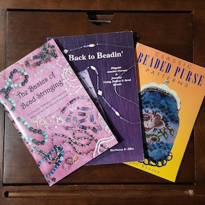 May include: Three beading books with colorful covers. The first book is pink and features the title "The Basics of Bead Stringing". The second book is purple and features the title "Back to Beadin'" with the subtitle "Elegant Amulet Purses & Jewelry Using Delica & Seed Beads". The third book is orange and features the title "Classic Beaded Purse Patterns".