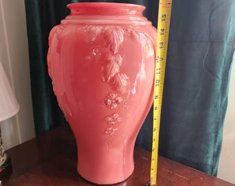 Op de afbeelding: Een grote, koraalkleurige glazen vaas met reliëf bloemen- en bladerenontwerpen. De vaas is ongeveer 40 cm hoog. De vaas heeft een brede body en een smallere hals. De vaas staat op een houten oppervlak.