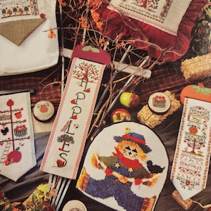 May include: A collection of autumn-themed cross-stitch projects. The designs include a scarecrow, apple trees, and the text "Apples" and "Autumn Harvest". The color palette features red, orange, green, and brown hues, evoking a fall harvest theme.