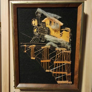 May include: A framed artwork depicting a train on a bridge. The train and bridge are constructed with gold-colored wire and wood, set against a black background. Fluffy white material suggests smoke. The frame is a warm brown.