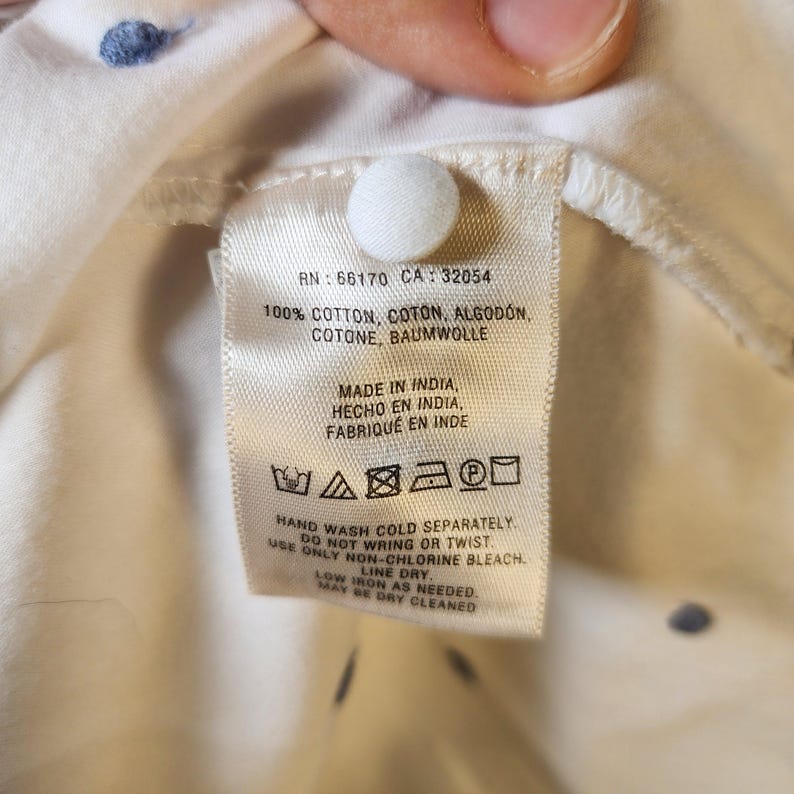 May include: White cotton shirt tag with care instructions. The tag includes the following information: RN: 66170 CA: 32054, 100% Cotton, Coton, Algod&oacute;n, Cotone, Baumwolle, Made in India, Hecho en India, Fabrique en Inde, Hand wash cold separately, Do not wring or twist, Use only non-chlorine bleach, Line dry, Low iron as needed, May be dry cleaned.