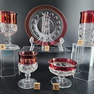 May include: Five pieces of vintage glass tableware with a ruby red rim. The set includes a plate, two goblets, a wine glass, and a small bowl.
