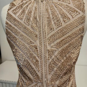 May include: A sleeveless bodice made of sheer nude mesh fabric, densely embellished with metallic gold glass beads arranged in a geometric, radiating pattern that extends from the neckline down across the chest and torso area.