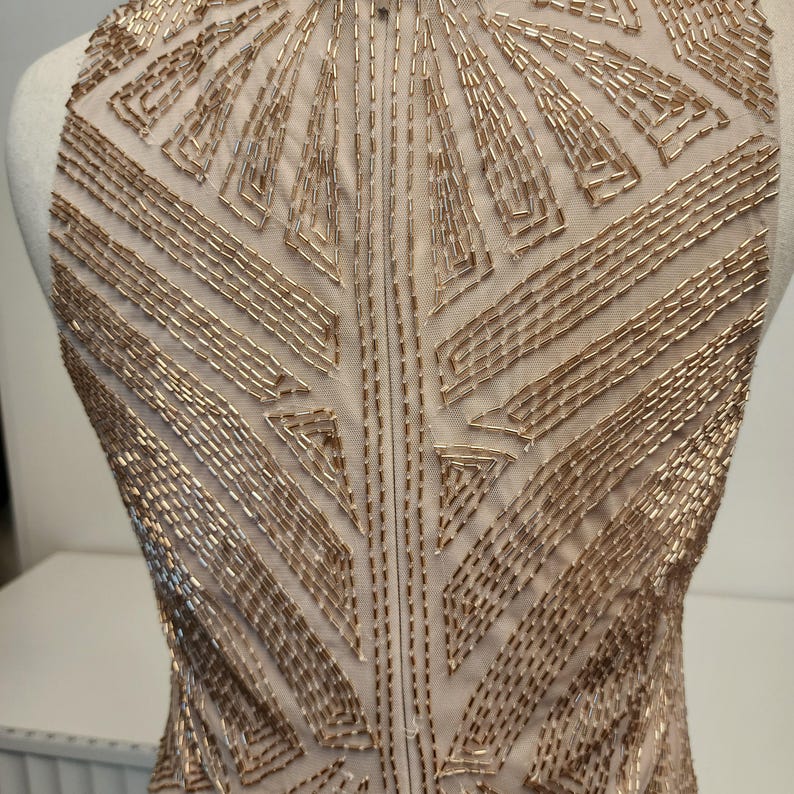 May include: A sleeveless bodice made of sheer nude mesh fabric, densely embellished with metallic gold glass beads arranged in a geometric, radiating pattern that extends from the neckline down across the chest and torso area.