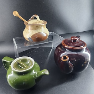 May include: Three ceramic teapots, one green, one brown, and one brown and beige with a wooden lid and handle. The teapots are on a clear glass surface.