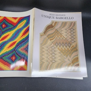 May include: Open book with colorful needlepoint patterns. The left page features a vibrant rainbow design, while the right page showcases a beige and brown Bargello pattern. The book's title reads "Jean Hilton's Unique Bargello."