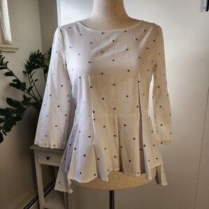 May include: A white, long-sleeved blouse with a white background and small blue polka dots. The blouse has a slightly flared hemline and a high, round neckline.