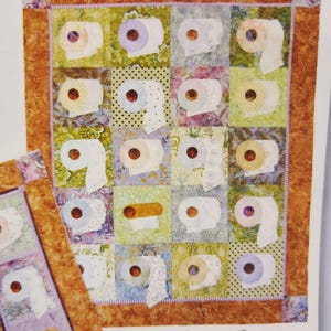 May include: A handmade quilt featuring squares with appliqued toilet paper rolls in various colors and patterns. The quilt has a brown border and a smaller matching piece is visible.