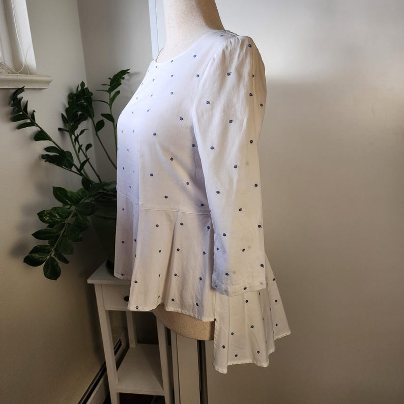 May include: White blouse with a blue polka dot pattern. The blouse has a high neckline and a high-low hemline. The sleeves are three-quarter length.