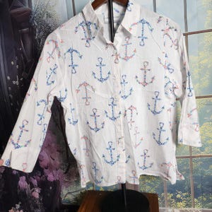 May include: White button-down shirt with a pattern of blue and pink floral anchors. The long-sleeved shirt has a collared neckline and a single pocket. The fabric has a subtle vertical stripe texture.