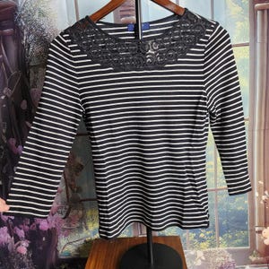 May include: Black and white striped long-sleeve shirt with a dark lace collar. The shirt has a scoop neck and three-quarter sleeves. The shirt is hanging on a wooden hanger.