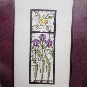 May include: Cross-stitch pattern featuring a floral design. The top section has a yellow rose surrounded by green leaves. The lower section displays purple irises with green stems and leaves, all within a dark brown frame.