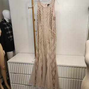 May include: A floor-length sleeveless dress in a light champagne color, featuring an intricate geometric pattern of gold sequins and beads throughout the bodice and skirt, with a flared hemline made of sheer tulle fabric and a round neckline.
