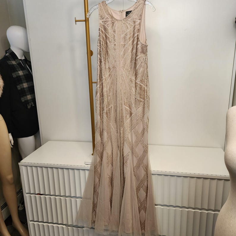 May include: A floor-length sleeveless dress in a light champagne color, featuring an intricate geometric pattern of gold sequins and beads throughout the bodice and skirt, with a flared hemline made of sheer tulle fabric and a round neckline.