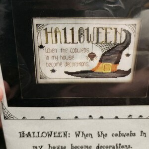 May include: Framed Halloween cross-stitch with the text "HALLOWEEN: When the cobwebs in my house become decorations." The design features a witch's hat, spiderwebs, and spiders in black, orange, and beige.