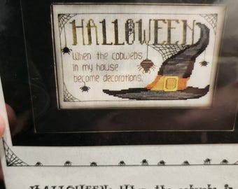 May include: Framed Halloween cross-stitch with the text "HALLOWEEN: When the cobwebs in my house become decorations." The design features a witch's hat, spiderwebs, and spiders in black, orange, and beige.