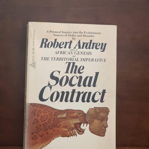 May include: A book cover with a white background and black text. The title is "The Social Contract" by Robert Ardrey, author of "African Genesis" and "The Territorial Imperative". The cover features an illustration of a leopard biting the head of a human.