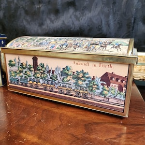 May include: Rectangular tin box with a hinged lid. The box features a detailed illustration of a town scene with horse-drawn carriages and the text "Ankunft in Fürth." The lid has a similar illustration of people on horseback. The box has a gold-colored trim.