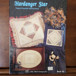 May include: A book titled "Hardanger Star" with teach-yourself instructions for embroidery. The cover features three examples of hardanger embroidery: a pillow, a small piece of fabric, and a piece of fabric in an oval frame. The book is by June Origo/designs Inc. and is book number 32.
