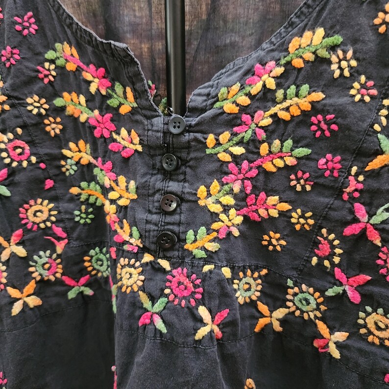 Peut inclure: Haut noir brod&eacute; avec un d&eacute;collet&eacute; en V et une fermeture boutonn&eacute;e. Le haut pr&eacute;sente des broderies florales color&eacute;es dans les tons de rose, jaune, orange et vert. La broderie est d&eacute;taill&eacute;e et couvre le devant du haut.