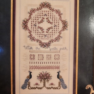 May include: A framed cross-stitch sampler with a floral design and the text "Walk the gentle path." The sampler features a central floral motif, geometric patterns, and two peacocks flanking a floral arrangement. The color palette includes shades of pink, blue, and cream.