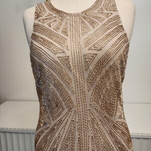 May include: A sleeveless dress featuring a beige base fabric adorned with intricate gold beadwork arranged in geometric patterns. The bodice displays a high neckline and a structured design, with the metallic beads creating symmetrical lines.