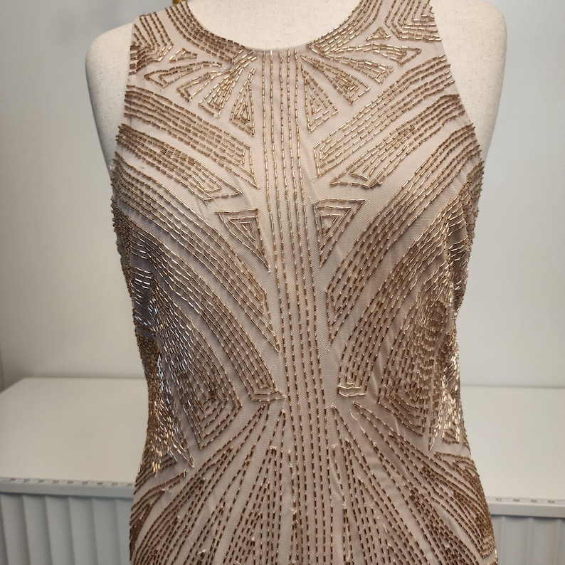 May include: A sleeveless dress featuring a beige base fabric adorned with intricate gold beadwork arranged in geometric patterns. The bodice displays a high neckline and a structured design, with the metallic beads creating symmetrical lines.