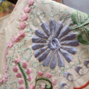 May include: Close-up of a cream-colored fabric with intricate embroidery. The design features a large, light blue flower in the center, surrounded by pink and green floral patterns. The stitching is detailed and adds texture to the fabric.