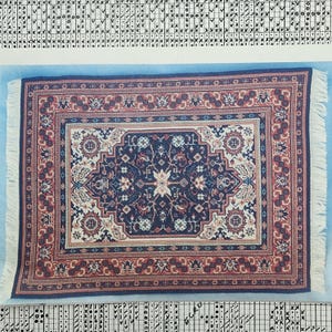 Puede incluir: Una alfombra rectangular con un campo central azul marino con un diseño floral en blanco y beige. La alfombra tiene un borde rojo con patrones intrincados y flecos blancos. Un elemento decorativo para el hogar.