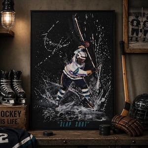 May include: A framed painting of a hockey player in action, titled "SLAP SHOT." The artwork features a player in a white, blue, and purple uniform, mid-slap shot, with white paint splatter effects. Hockey equipment, including skates and a helmet, is visible.