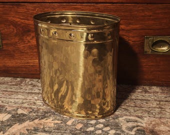 May include: An oval-shaped brass wastebasket featuring a hammered texture on the body and a smooth upper band decorated with a series of evenly spaced circular indentations around the entire circumference of the metal container.