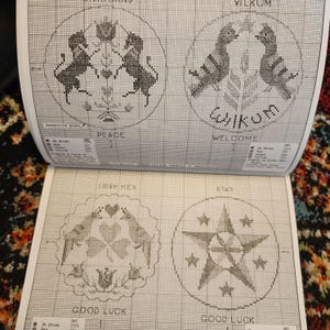 May include: A page from a cross-stitch pattern book featuring four circular designs. The designs include unicorns, birds, and stars, with text such as "Peace," "Welcome," and "Good Luck." The patterns are in black and white.